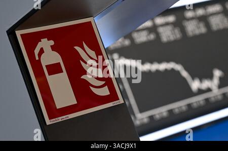 04 April 2025, Hesse, Frankfurt/Main: A sign for a fire extinguisher is displayed at a passageway of the Frankfurt Stock Exchange, while the display board with the Dax curve shows falling prices. US President Trump had issued a huge tariff package against trading partners around the world. The European Union and China have already announced countermeasures. Photo: Arne Dedert/dpa Stock Photo