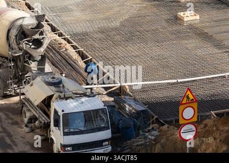A construction site is active, featuring a concrete mixer truck pouring cement onto newly laid steel rebar. A worker stands nearby, ensuring the proce Stock Photo