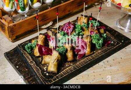 Appetizer platter featuring stuffed eggplant rolls garnished with kale and radicchio, served on rustic wooden tray with skewers. Stock Photo