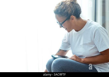 Woman sitting and using her phone to text and read notifications, browsing social media, checking emails, smiling while interacting with her smartphone, and texting with friends or family members Stock Photo