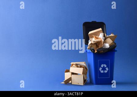 Blue trash container with recycling symbol filled with cardboard and paper waste and and other boxes and paper next to it on the ground isolated with Stock Photo