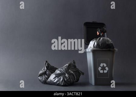 Gray trash container with recycling symbol filled with garbage bag and other bags next to it on the ground isolated with gray gradient background. Fro Stock Photo