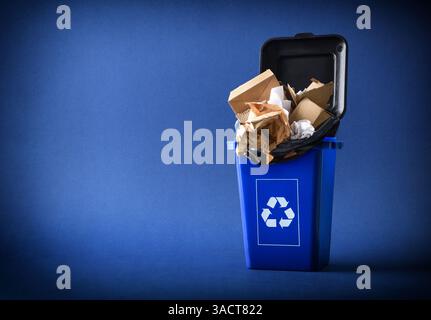 Cardboard and paper recycling concept with a blue bin with the recycling symbol and a black bag isolated on a blue background. Front view. Stock Photo