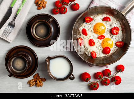 Two fried eggs with cherry tomatoes in a pan, two cups of coffee, milk, nuts and cherry tomatoes branches are composing a delicious breakfast on a white wooden table Stock Photo