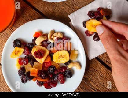 Hand picking crispy dried banana chips from a colorful mix of nuts and dried fruits, beautifully arranged on a white plate atop a wooden table, accompanied by a refreshing glass of fruit juice Stock Photo