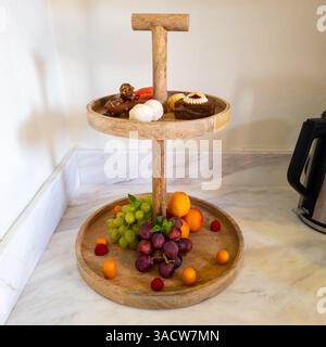 Two-tiered wooden stand showcasing an assortment of fresh fruits, pastries, and sweets on marble surface. Stock Photo