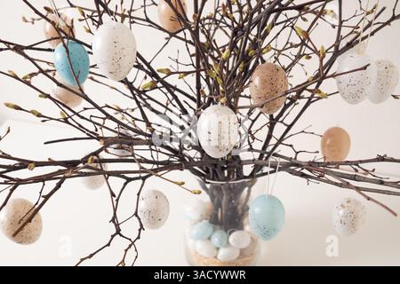 Easter tree decorated colorful pastel white and beige eggs. Birch branches with hang eggs in dark vase on white table. Close up Stock Photo
