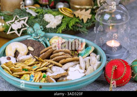 Plate with various Christmas cookies, arranged with a glass lantern and Christmas baubles in front of an Advent wreath on a table runner Stock Photo