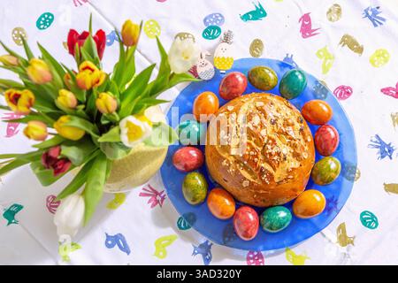 Easter cake with colorful Easter eggs on a blue glass plate, tablecloth with Easter motifs and tulips Stock Photo