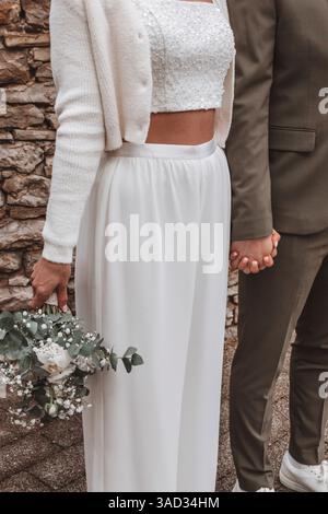 Bride and groom standing next to each other, bride wearing a belly-baring pantsuit instead of a dress in the registry office, couple holding hands Stock Photo