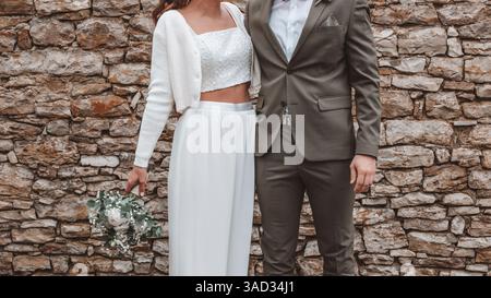 Bride and groom standing next to each other, bride wearing a belly-baring pantsuit instead of a dress in the registry office, couple holding hands Stock Photo