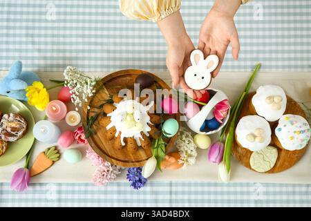 Female hands holding gingerbread in shape of bunny near table setting for Easter celebration. Top view Stock Photo