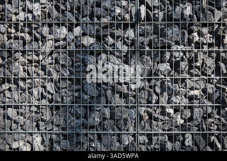 A tightly packed gabion wall consisting of rough gray rocks held together in a structured metal wire grid. Stock Photo