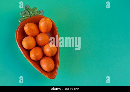 Minimalist Easter-themed arrangement featuring bright chocolate eggs in a ceramic carrot dish on a pastel background Stock Photo