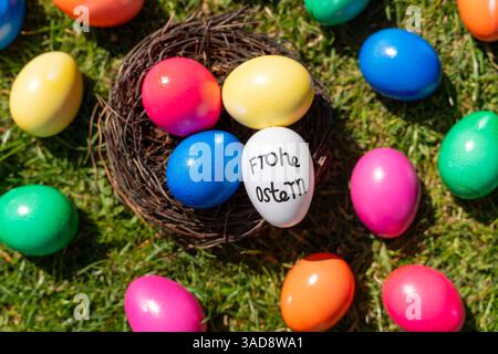 Bavaria, Germany - 5 April 2025: Colorful Easter eggs lie in a nest on green grass. A white egg with the inscription Happy Easter combines festive decoration with warm Easter wishes and the joy of spring. *** Bunte Ostereier liegen in einem Nest auf grünem Gras. Ein weißes Ei mit der Aufschrift Frohe Ostern verbindet festliche Dekoration mit herzlichen Osterwünschen und Frühlingsfreude. Stock Photo