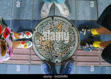 Nara, Japan - Sep 26 2024, A close-up view of a sewer manhole patterned with a colored image of a deer among foliage and trees, surrounded by the legs Stock Photo