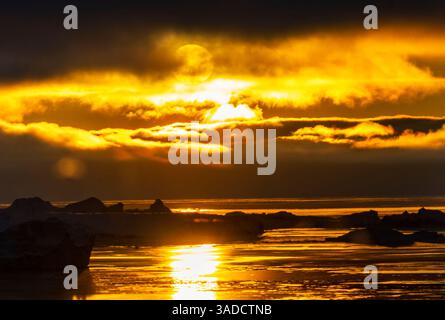 A stunning sunset over icy waters, with vibrant orange and yellow hues reflecting off the surface. Icebergs are visible in the foreground, creating a Stock Photo