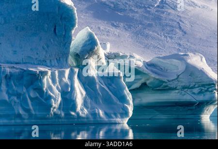 A close-up view of an iceberg with intricate textures and shapes, reflecting in calm waters. The surrounding landscape features snow-covered terrain u Stock Photo