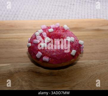 A bright pink donut garnished with white and pink marshmallows sits on a wooden cutting board. The warm kitchen ambiance enhances the appeal of this s Stock Photo