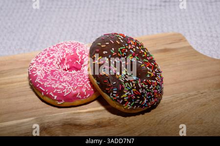 Two delectable donuts sit on a wooden board, one coated with pink icing and white sprinkles, and the other with a rich chocolate topping and vibrant m Stock Photo