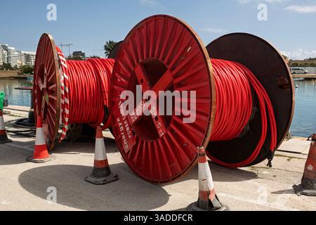 Large red industrial cable reels on a port dock with a sea bay in the background, surrounded by plastic safety cones. Concept of urban construction Stock Photo