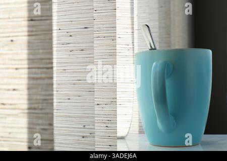 A blue mug with a spoon stands in front of blinds, with soft sunlight gently filtering through. Stock Photo