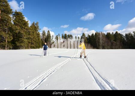Skiers take break in snowy field, person in yellow looking down at gear while companion with poles stands still, enjoying scenery on bright winter day Stock Photo