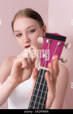 Young woman musician holding her ukulele and tuning strings of musical instrument, tightening and loosening strings before beginning to play ukulele. Stock Photo