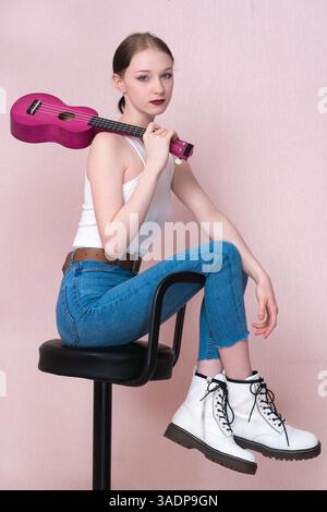 Young female musician sitting on bar stool, holding ukulele by neck and throwing musical instrument over her shoulder, looking at camera. Stylish hips Stock Photo