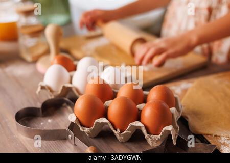 Close-up of brown and white eggs in carton with child rolling dough in cozy kitchen, baking preparation scene. Stock Photo