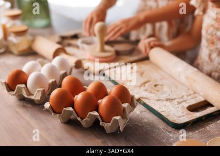 Children baking together with fresh eggs, rolling dough and cutting shapes on a floured wooden kitchen surface. Stock Photo