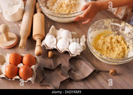 Children whisk dough in glass bowls surrounded by fresh eggs, baking tools, and flour on a wooden table. Stock Photo