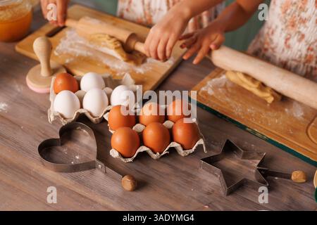 Children rolling out cookie dough on wooden boards with eggs and metal cutters during home baking fun. Stock Photo