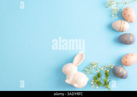 White ceramic bunny with pastel decorated easter eggs and flowers on blue background. Easter holiday concept. Top view, flat lay, copy space. Stock Photo