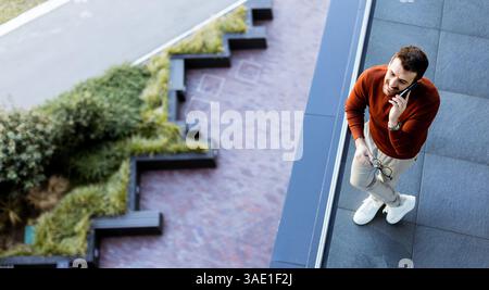 A young man stands on a sleek balcony while engaged in an animated phone conversation. Below him, a blend of contemporary design and lush greenery enh Stock Photo