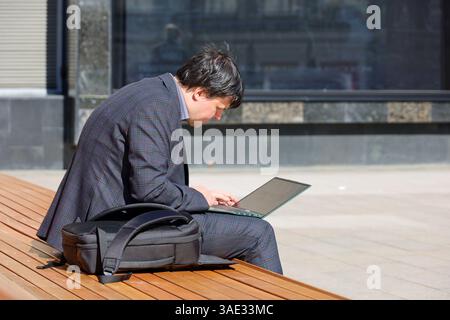 Man in a business suit working with laptop on a bench on city street Stock Photo
