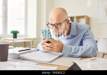 Funny young businessman using smart phone watching screen at desk, smiling male professional  Stock Photo