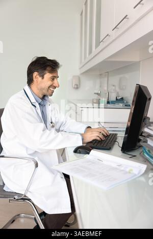 Smiling Latin male doctor working at desk using computer Stock Photo