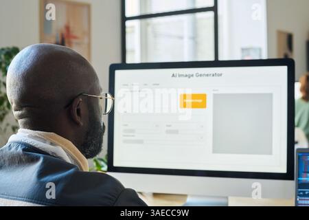 Focus on bald head of young African American designer sitting in front of desktop computer monitor and using AI image generator Stock Photo