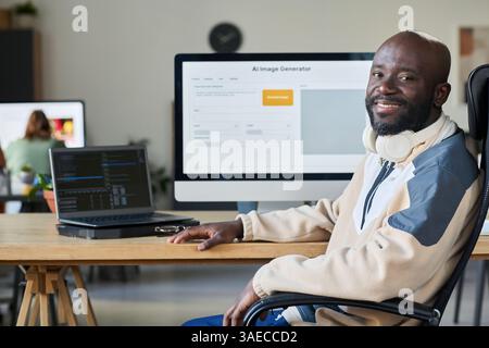 Young successful African American male programmer looking at camera while sitting by workplace with two computers Stock Photo