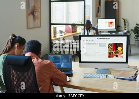 Rear view of two young intercultural programmers or designers sitting by workplace with desktop computer with image generator on screen Stock Photo