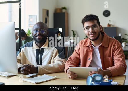 Two happy young intercultural male programmers looking at camera while using desktop computer and tablet in coworking space Stock Photo