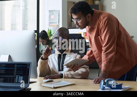 Serious young man with tablet bending over desk while standing next to colleague and both looking at computer screen Stock Photo