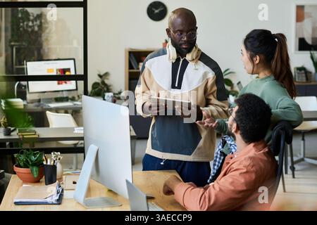 Young serious male programmer with tablet looking at Chinese female colleague during discussion of new project at meeting Stock Photo