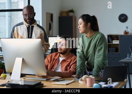Three intercultural designers or programmers looking at new software product on computer screen in coworking space Stock Photo