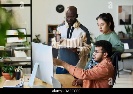 Group of young colleagues looking at screen of desktop computer while one of them pointing at new software product Stock Photo