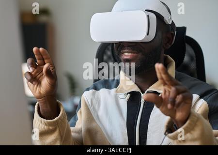 Young smiling African American man scrolling through virtual presentation or information in VR headset while sitting in office Stock Photo