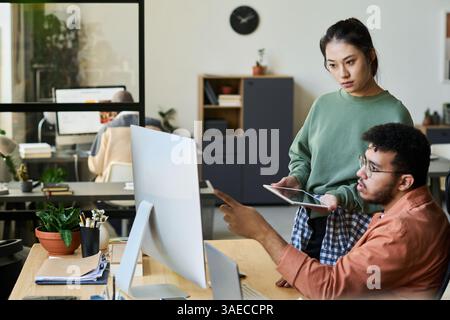 Young male programmer pointing at computer screen while discussing ways of decoding data with female colleague at meeting Stock Photo