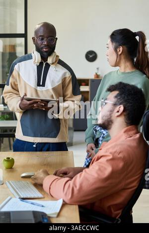Young African American programmer with tablet in hands looking at his colleagues during discussion of new software Stock Photo