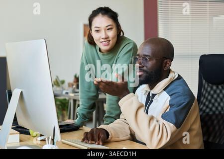 Happy young Asian female programmer standing next to male colleague explaining her points of new project by workplace Stock Photo